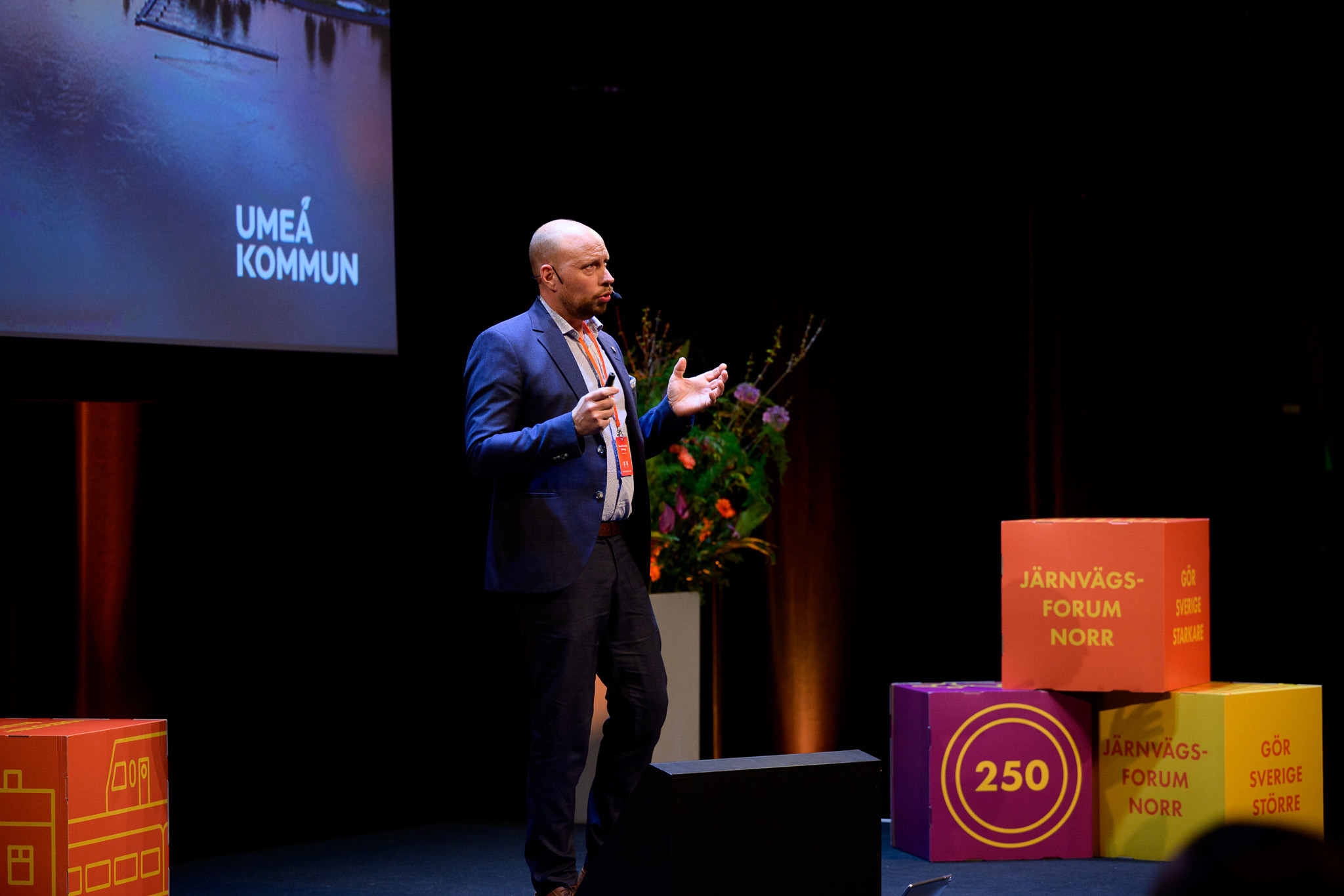 En man står ensam på scen och talar under Järnvägsforum Norr. På storskärmen bakom honom syns texten ”Umeå kommun”. Till höger står färgglada budskapskuber med ord som ”250”, ”Järnvägsforum Norr” och ”Gör Sverige starkare”.