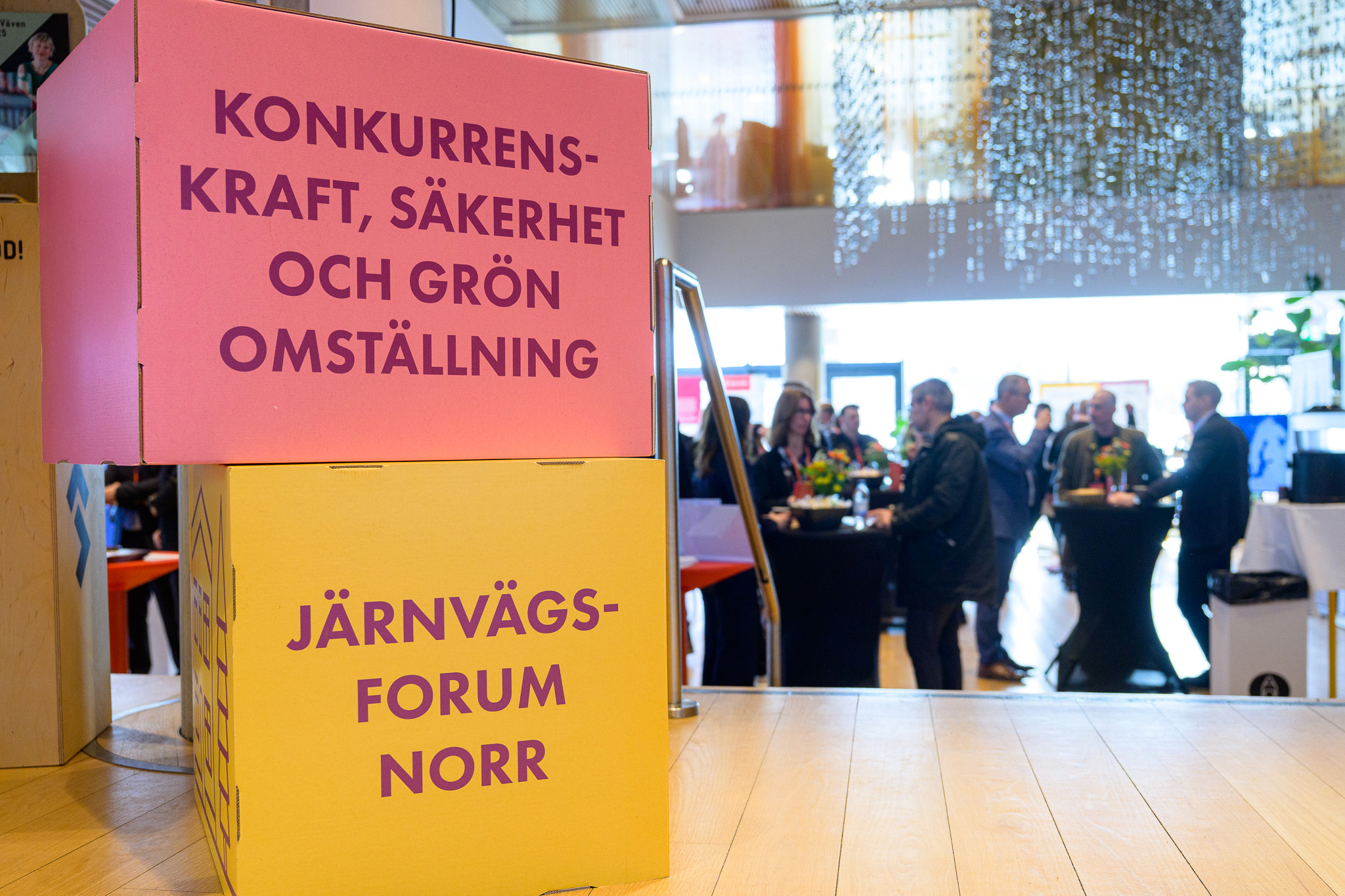 Två staplade kuber med budskapet ”Konkurrenskraft, säkerhet och grön omställning” samt ”Järnvägsforum Norr” står i förgrunden. I bakgrunden syns ett mingelområde med ståbord och deltagare i samtal under ett glittrande tak i en ljus lokal.