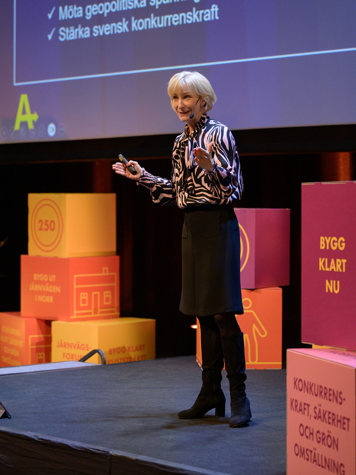 En kvinna står på scen och föreläser under Järnvägsforum Norr. Hon har mikrofon och fjärrkontroll i handen, och bakom henne visas en presentation med punkter som ”Stärka svensk konkurrenskraft”. Runt scenen syns färgglada kuber med budskap som ”Bygg klart nu” och ”Konkurrenskraft, säkerhet och grön omställning”.