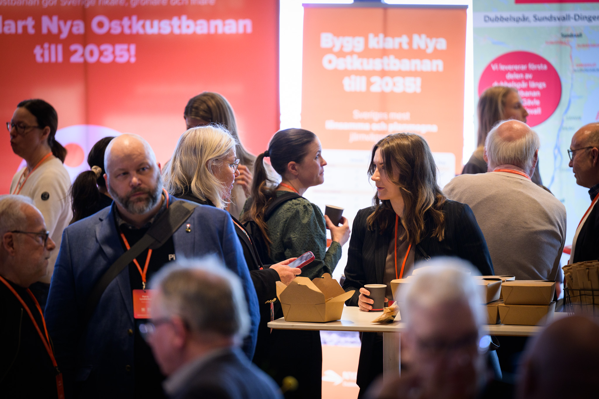 Personer minglar och samtalar vid ett ståbord under Järnvägsforum Norr. I förgrunden syns matlådor och kaffemuggar, i bakgrunden stora skyltar med texten ”Bygg klart Nya Ostkustbanan till 2035!”.